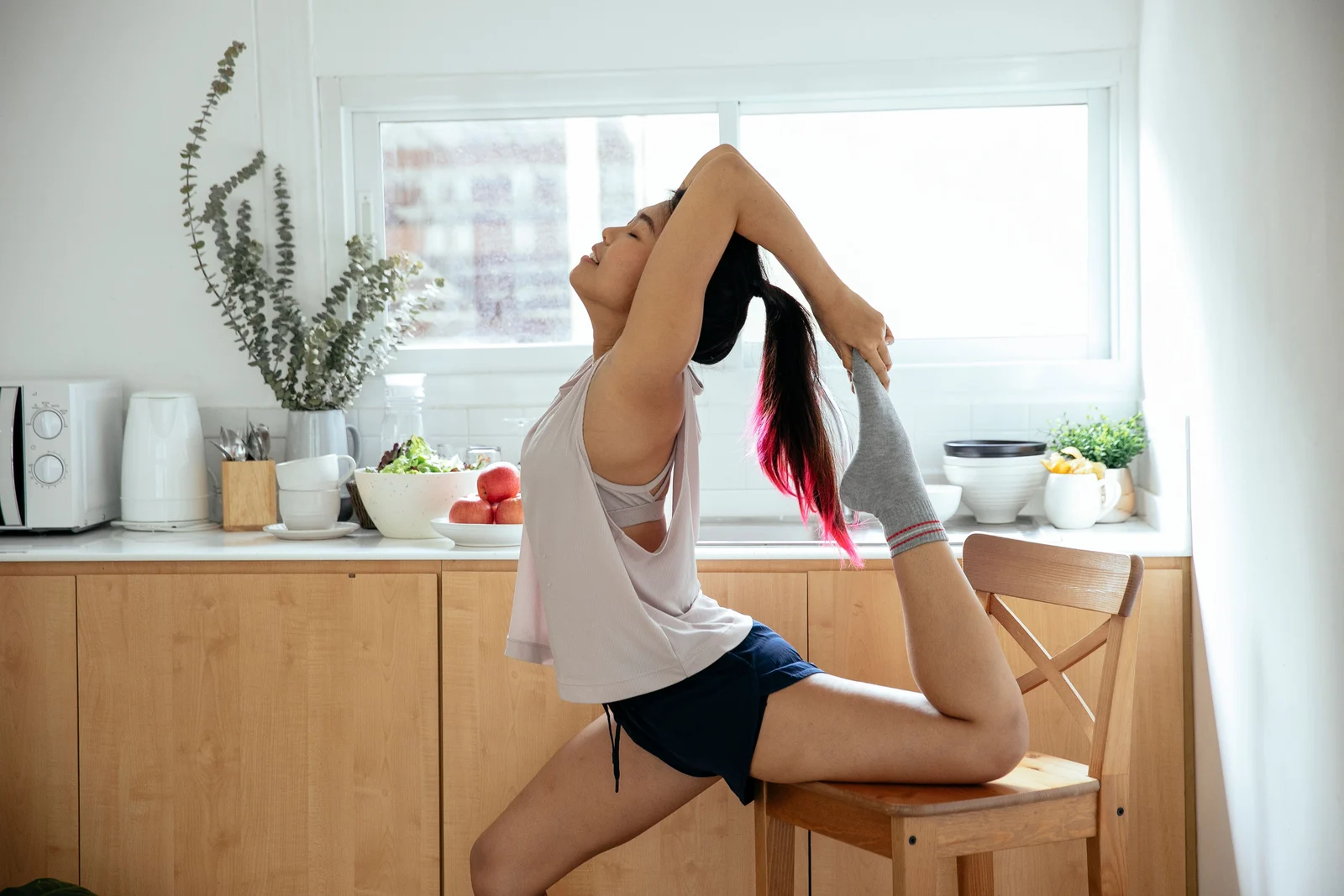 A woman practising yoga at home, using a kitchen chair to support a standing stretch.