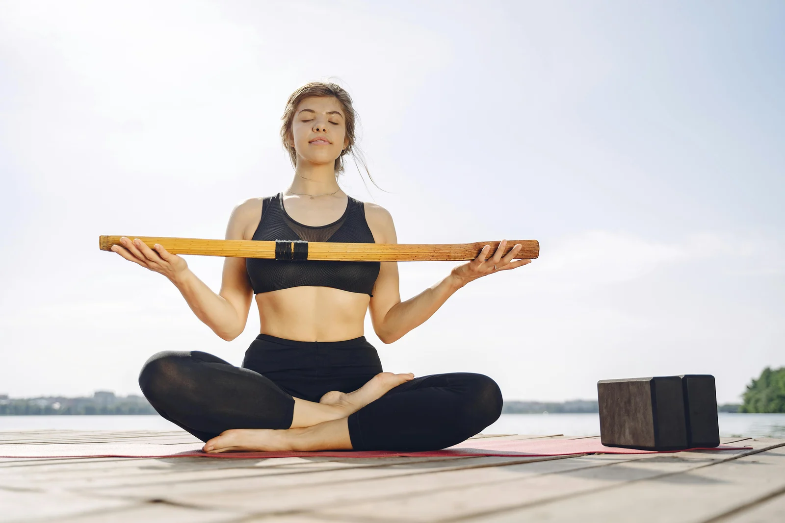 A woman sitting on a yoga mat by a lake, yoga blocks resting on the mat beside her.